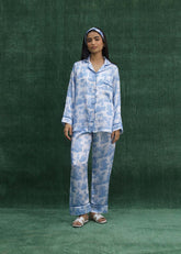 A woman standing against a green background, wearing a white silk pajama set with powder blue leaf prints, consisting of a button-down shirt with front pockets and trousers with lined hems.